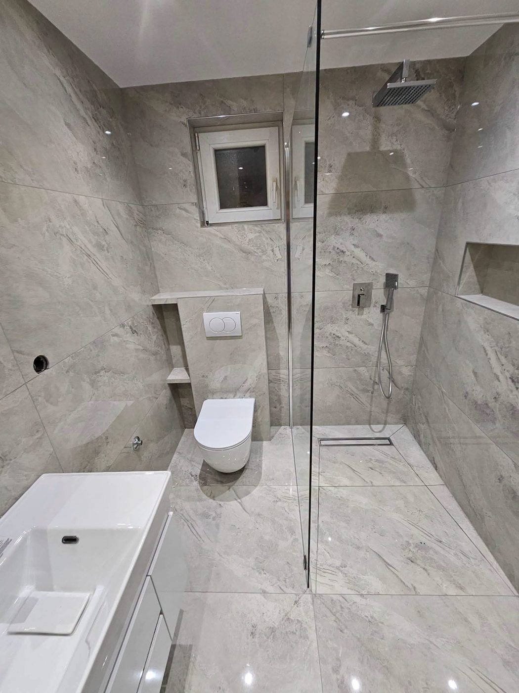 Finished bathroom with large grey stone-effect tiles, a wall-hung toilet and a glass shower screen